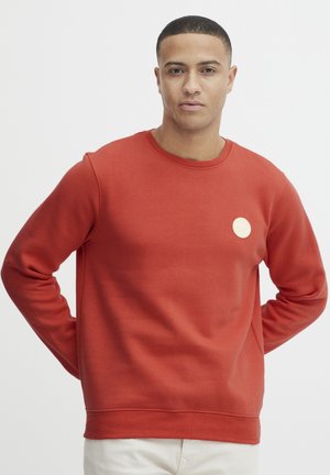 Sweatshirt roșu cu guler circular, fabricat dintr-un material moale, având un guler ribat și un mic logo texturat pe partea stângă a pieptului.