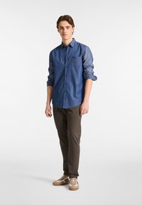 Denim overhemd met een knoopsluiting aan de voorkant, een borstzak en opgerolde mouwen, gecombineerd met donkerbruine broek en beige sneakers met lichte accenten.