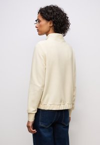Felpa con zip color crema, caratterizzata da un motivo testurizzato, polsini e vita a coste, indossata con jeans blu scuro. Vista da dietro.
