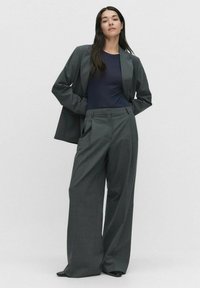 Harmaan räätälöidyn puvun yläosana pitkä bleiseri, jossa on läpät, ja leveälahkeiset housut, yhdistettynä istuvaan tummansiniseen yläosaan. Sileä rakenne, ryhdikäs istuvuus.