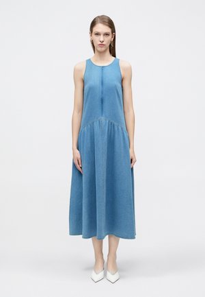 Femme portant une robe midi en denim bleu sans manches et des talons blancs pointus, debout devant un fond uni clair.