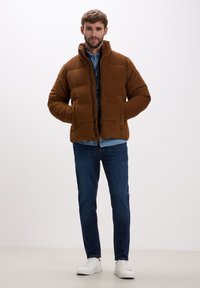 Giacca a pirata marrone con colletto alto, chiusura frontale con zip e due tasche laterali; indossata sopra una camicia di denim e abbinata a jeans scuri e sneaker bianche.