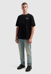Zwarte katoenen T-shirt met korte mouwen en een ronde hals, voorzien van een kleine graphic op de borst; gecombineerd met versleten lichtblauwe jeans en zwarte schoenen.