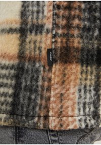 Kurtka w kratę z połączeniem kremowego, brązowego i szarego koloru, wykonana z miękkiego, puszystego materiału. Czarna metka z napisem "CHASIN'" blisko szwu.