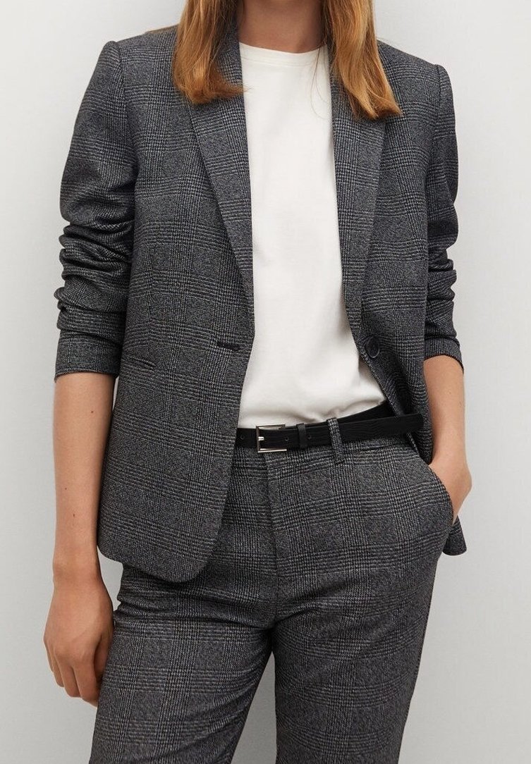 Femme portant un blazer gris à carreaux et un pantalon assorti avec une ceinture noire et un haut blanc, une main dans la poche, debout devant un fond uni.