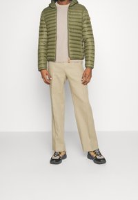 Giubbotto imbottito verde oliva con cappuccio, maglione a maglia beige, pantaloni larghi beige chiaro e sneakers nere e gialle con lacci.