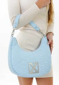 Femme portant une robe crème côtelée tient un sac à bandoulière matelassé bleu clair avec un emblème géométrique argenté et une large sangle.