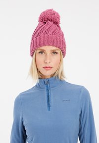 Berretto lavorato a maglia rosa con un grande pompon sopra, caratterizzato da una struttura a coste spesse e motivi diagonali. Indossato su un top in pile blu.