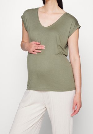 Kvinde iført olivengrøn kortærmet top og lyse beige bukser, der blidt holder sin gravide mave med en hånd.