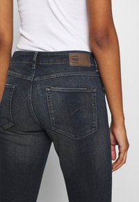 Tmavě modré denimové džínsy s klasickým střihem, vybavené hnědou koženou nášivkou a dvěma zadními kapsami, viditelným švem a minimálním vyblednutím.