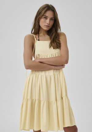 Giovane donna con lunghi capelli castani che indossa un vestito giallo pallido a balze senza maniche, in piedi con le braccia incrociate davanti a uno sfondo semplice.