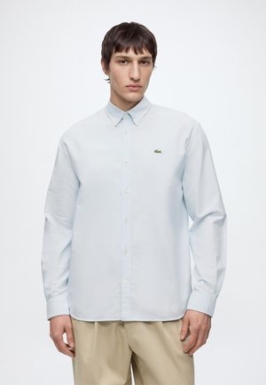 Jeune homme aux cheveux courts et foncés portant une chemise bleu clair boutonnée et un pantalon beige, debout devant un fond blanc uni.