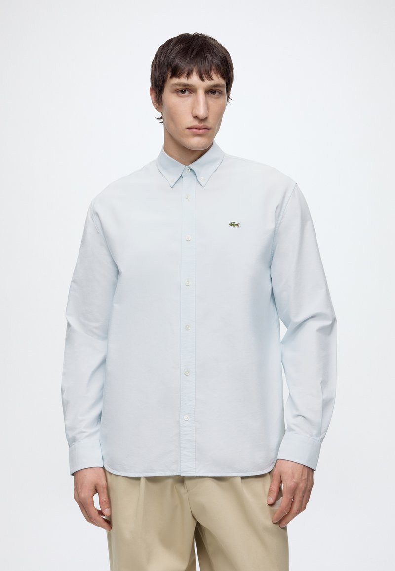 Giovane uomo con capelli corti e scuri che indossa una camicia azzurro chiaro con bottoni e pantaloni beige, in piedi davanti a uno sfondo bianco semplice.