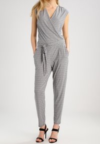Jumpsuit à motif noir et blanc avec des manches courtes, design en wrap, ceinture à la taille et poches latérales. Accompagné de talons noirs.