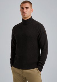 Pull à col roulé marron foncé avec un design en mailles texturé, des poignets côtelés et une silhouette ajustée. Comprend un petit patch de marque sur la manche.