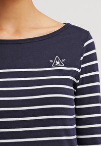 Navy blue shirt with white horizontal stripes and a small white embroidered triangular emblem with "1897" near the neckline.