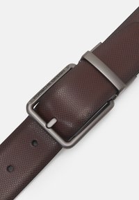 Cinturón de cuero marrón con patrón y hebilla de metal cepillado. Inscripción "Porsche Design" en la hebilla, con orificios distintivos para ajustes.