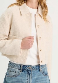 Veste beige texturée et courte avec un col, comportant cinq grands boutons et une poche poitrine, portée sur une chemise blanche.