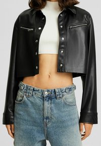 Czarna skórzana kurtka o krótkim kroju z zamkami błyskawicznymi w kolorze srebrnym, z guzikami oraz długimi rękawami, zestawiona z wyblakłymi niebieskimi dżinsami.
