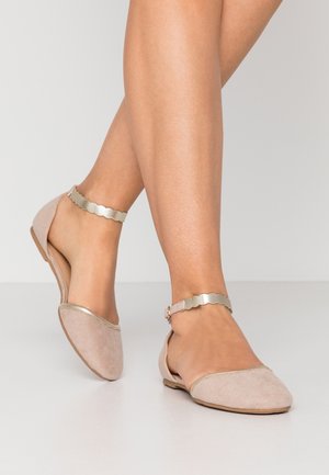 Piernas que llevan zapatos planos beige con correas metálicas en el tobillo con ondas y puntera cerrada, de pie sobre un suelo gris claro.