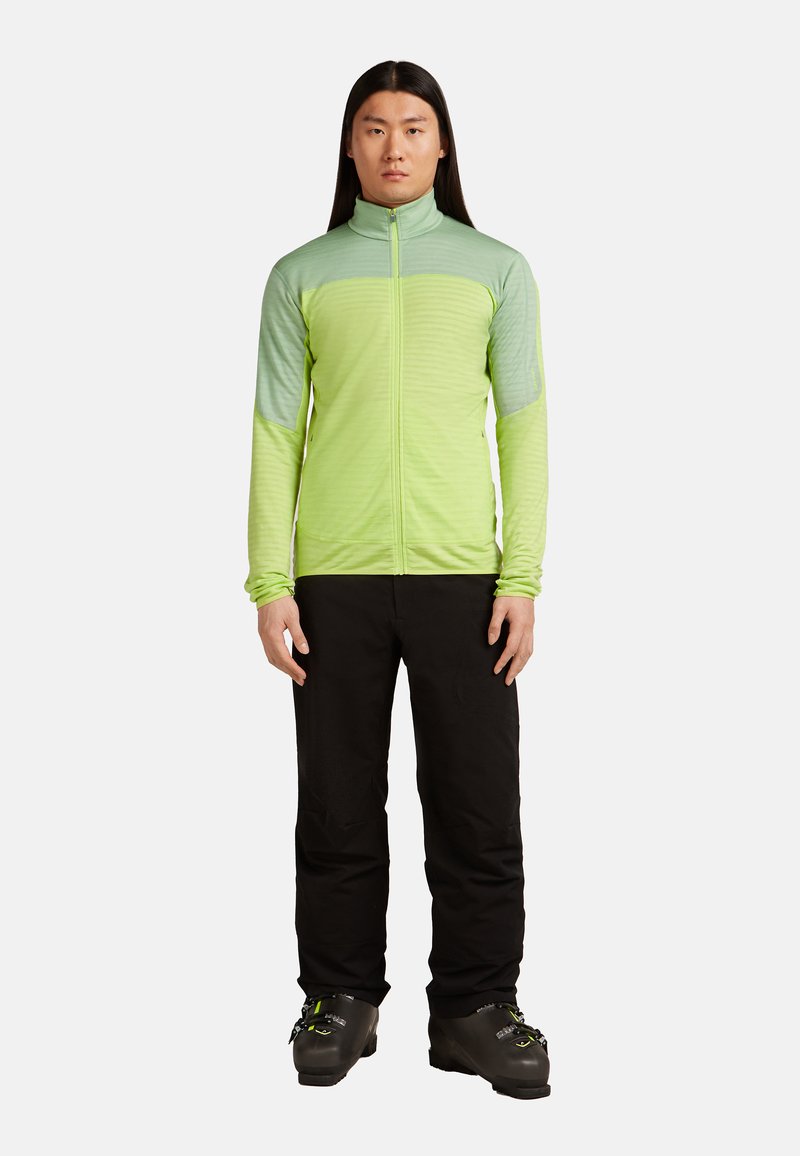 Hombre con cabello largo y negro que lleva una chaqueta verde con cremallera, pantalones negros y botas de esquí negras, de pie contra un fondo blanco.