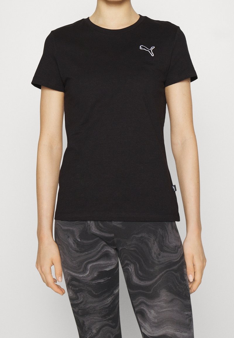 Femme portant un t-shirt noir ajusté Puma avec un logo blanc et des leggings noirs et gris à motifs, debout sur un fond uni.