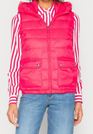 Vrouw draagt een felroze doorgestikte capuchonvest over een rood-wit gestreept overhemd met knopen en blauwe spijkerbroek.