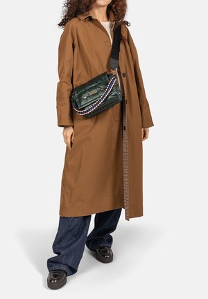 Gewatteerde groene handtas met een kettingaccent, als crossbody gedragen. Gecombineerd met een bruine jas, blauwe spijkerbroek en zwarte loafers. Soepele textuur.