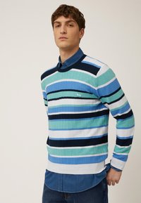 Maglione lavorato a maglia con righe orizzontali nelle tonalità di blu, verde e navy; texture a costine; scollatura a girocollo; indossato sopra una camicia di denim blu.