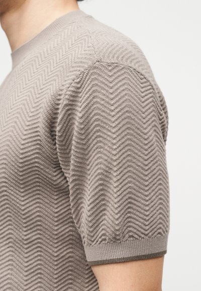 Κοντινό πλάνο ενός ατόμου που φοράει μπεζ πλεκτό τοπ με κοντά μανίκια και υφή ζιγκ-ζαγκ σε απλό φόντο.