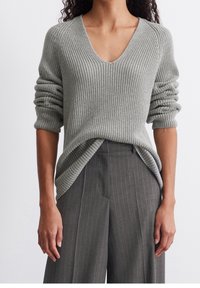 Femme portant un pull gris côtelé à col en V avec des manches retroussées et un pantalon à jambes larges taille haute gris à fines rayures.