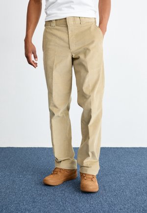 Beige kordflöjlsbyxor med rak passform. Har framfickor och dragkedja. Matchade med bruna sneakers på en blå matta.