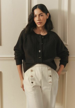 Frau mit dunklem, welligem Haar, die einen schwarzen, geknöpften Pullover und hochgeschnittene, cremeweiße Hose trägt, steht vor einer cremefarbenen Wand.