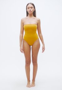 Ολόσωμο μαγιό χωρίς τιράντες σε μουσταρδί κίτρινο χρώμα, με λεία υφή και ψηλές περικοπές στα πόδια, σχεδιασμένο για μια κομψή σιλουέτα.