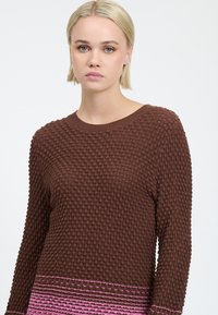 Jonge vrouw met kort blond haar, gekleed in een bruine gebreide trui met roze gestreepte details aan de onderkant, tegen een witte achtergrond.