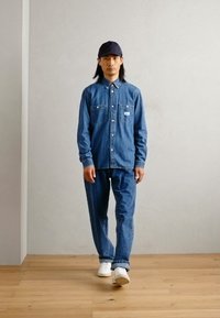 Denim shirt with button closure and two pockets, paired with matching denim pants and white sneakers. The individual wears a navy cap.