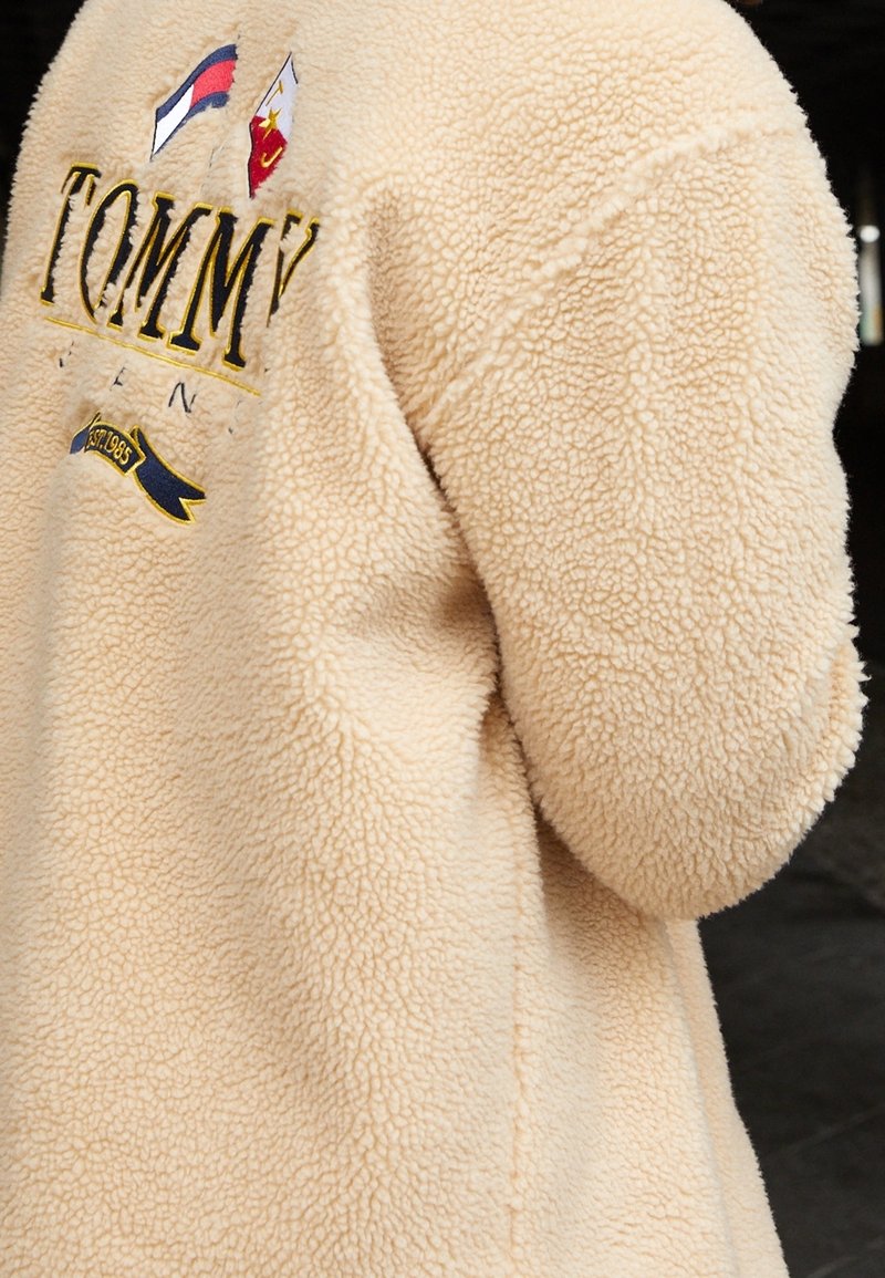 Veste en polaire beige duveteuse avec logo Tommy Hilfiger, drapeaux nautiques et broderie "Est. 1985" au dos.
