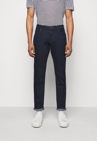 Osoba stoi w ciemnych dżinsach i w paskową koszulę, do których ma dopasowane białe sneakersy, na jednolitym tle.