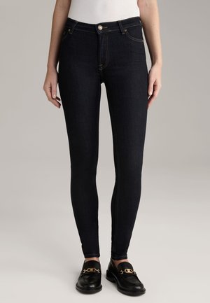 Kobieta nosząca czarne wąskie jeansy w połączeniu z czarnymi loafersami ze złotymi klamrami, stoi na jednolitym jasnym tle.