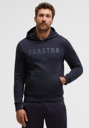 Mann trägt eine marineblaue Gaastra-Hoody mit Fronttasche, steht vor einem schlichten hellen Hintergrund und schaut in die Kamera.
