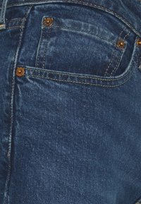 Los vaqueros de mezclilla presentan un color azul intenso con una textura desgastada. Hay dos botones de bronce en el bolsillo delantero y una costura amarilla que contrasta.