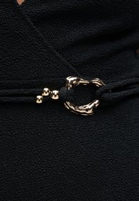 Κοντινό πλάνο μαύρου υφάσματος με υφή, με μαύρο κορδόνι που διαθέτει λαμπερή, ακανόνιστη χρυσή αγκράφα και τρεις μικρές χρυσές χάντρες.