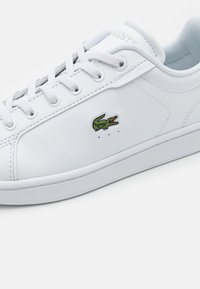 Hvit skinn sneaker med hvite snører og en liten grønn Lacoste krokodillelogo på sidepanelet.