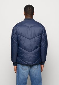 Marineblauwe donsjas met chevronste stitching, ribgebreide manchetten en een hoge kraag, ontworpen voor warmte en comfort.