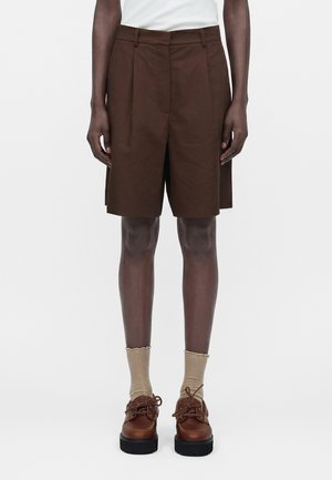 Άτομο που φοράει σκούρα καφέ σορτς έως το γόνατο, μπεζ κάλτσες και καφέ δερμάτινα παπούτσια με κορδόνια, στέκεται μπροστά σε απλό φόντο.