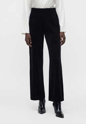 Femme portant un pantalon large en velours noir, une blouse blanche à manches longues et des bottes à talons noires à bout pointu, debout sur un fond blanc.