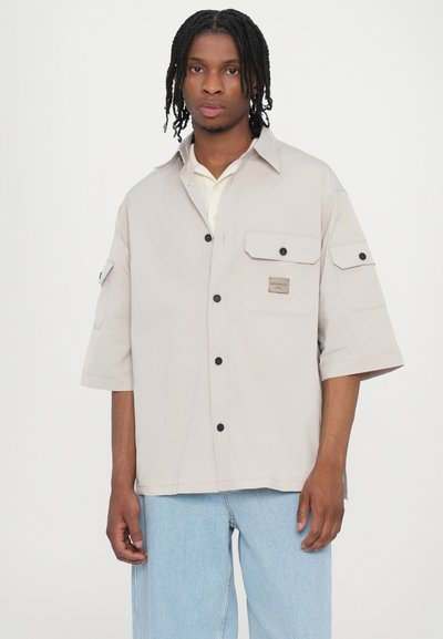 Chemise courte à manches dans un beige clair avec deux poches poitrine, boutons noirs et un patch logo, associée à un jean bleu clair ample.