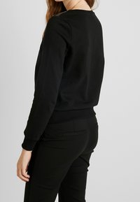 Sweatshirt noir à manches longues, poignets et ourlet côtelés. Texture lisse, encolure ronde, avec des détails de couture subtils aux épaules.