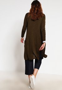 Długi kardigan w kolorze oliwkowej zieleni z długimi rękawami, kieszeniami i luźnym krojem, w połączeniu z ciemnymi, krótkimi jeansami i białymi trampkami.