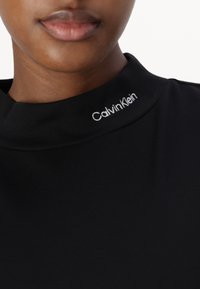 Melna augsta kakla drēbe ar mīkstu audumu. Balts "Calvin Klein" logos pie apkakles. Tuvplāns, kas parāda tekstūru un šuvju detaļas.